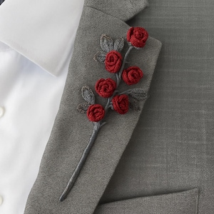 May include: A handcrafted red rose boutonniere on a gray blazer. The floral accessory features five red roses with gray leaves and stem. A white dress shirt is visible in the background. This elegant piece adds a touch of sophistication.
