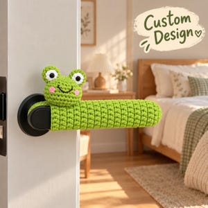 May include: A bright green crocheted frog door handle cover featuring a smiling frog face with black and white eyes and pink cheeks. The cover is fitted over a black door handle. The text Custom Design is visible in the upper right corner.