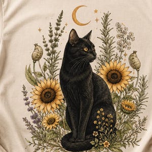 May include: A cream-colored t-shirt featuring a graphic print of a black cat sitting amidst a cluster of yellow sunflowers, lavender sprigs, and green foliage. Above the cat is a golden crescent moon with small star accents, all centered on the chest.