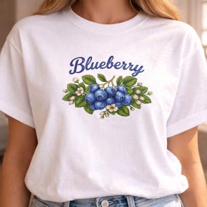 May include: White t-shirt with a colourful illustration of blueberries, green leaves, and small white flowers. The word "Blueberry" is written in dark blue script above the fruit design.
