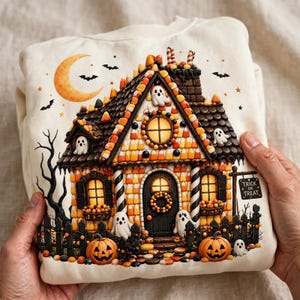 Op de afbeelding: Een crèmekleurig sweatshirt met een afbeelding van een huis gemaakt van candy corn met donkere chocolade dakpannen. De scène bevat verlichte ramen, kleine witte spookjes, oranje pompoenen, een maansikkel en een bordje met Trick or Treat.