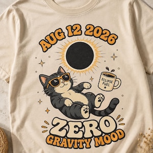 May include: A cream-coloured t-shirt featuring a graphic of a black and white cat wearing orange sunglasses, floating beneath a black sun. The text reads AUG 12 2026, ECLIPSE FUEL on a coffee mug, and ZERO GRAVITY MOOD, surrounded by small stars.
