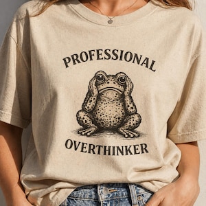May include: A beige t-shirt featuring the text "PROFESSIONAL OVERTHINKER" and a detailed illustration of a frog. The frog is brown with darker spots and is covering its eyes. The shirt has a crew neck and short sleeves.