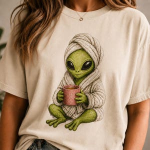 Könnte beinhalten: Beiges T-Shirt mit einer Illustration eines grünen Außerirdischen, der einen weißen Bademantel und eine Kopfbedeckung trägt und einen rosa Becher hält. Der Außerirdische hat große schwarze Augen und sitzt im Schneidersitz.
