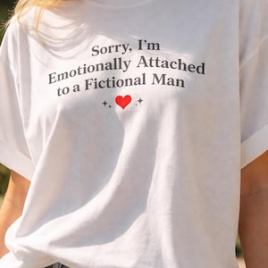 May include: White t-shirt with the text "Sorry, I'm Emotionally Attached to a Fictional Man" in dark gray, with a red heart and star accents. The shirt is paired with blue jeans.