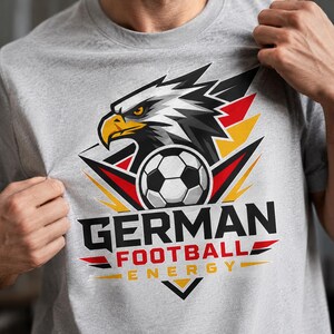 Op de afbeelding: Een grijs gemêleerd t-shirt met een afbeelding van een adelaarskop boven een voetbal. Het ontwerp bevat zwarte, rode en gele accenten en de tekst GERMAN FOOTBALL ENERGY in dikke zwarte en rode letters op de voorzijde van het kledingstuk.