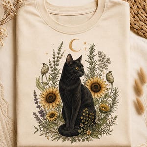May include: A cream-colored t-shirt featuring a graphic illustration of a black cat sitting amidst a cluster of sunflowers, lavender, and various botanical elements, topped with a golden crescent moon and small star accents in the center.