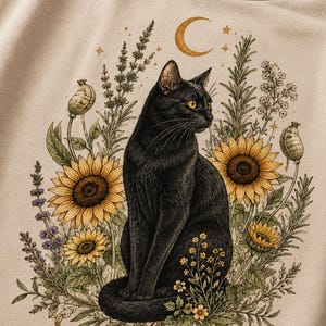May include: A beige t-shirt featuring a graphic print of a black cat sitting amidst sunflowers, lavender, and small yellow flowers. Above the cat is a golden crescent moon and stars. The illustration is detailed with botanical elements and foliage.