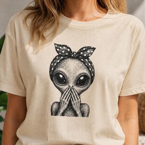 May include: Beige t-shirt with a black and white illustration of an alien. The alien has large eyes, hands covering its mouth, and a polka dot headband. The graphic is centered on the shirt.