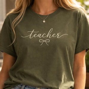May include: Olive green t-shirt with the word "teacher" in a cursive font and a small bow graphic underneath. The shirt is made of a soft material and is suitable for casual wear.