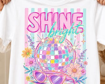 以下が含まれることがあります： ピンクとブルーの「SHINE bright」という文字がプリントされた白いTシャツ。デザインには、マルチカラーのミラーボール、ピンクのサングラス、様々な花の要素が描かれており、背景にはピンクとブルーの縦縞模様があしらわれています。