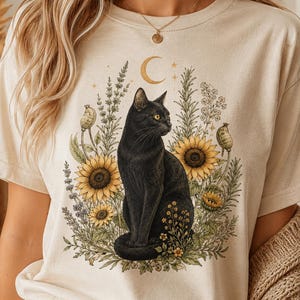 May include: A cream-colored t-shirt featuring a graphic print of a black cat sitting amidst yellow sunflowers, green foliage, and lavender sprigs, topped with a golden crescent moon and small stars, rendered in a detailed, illustrative style.