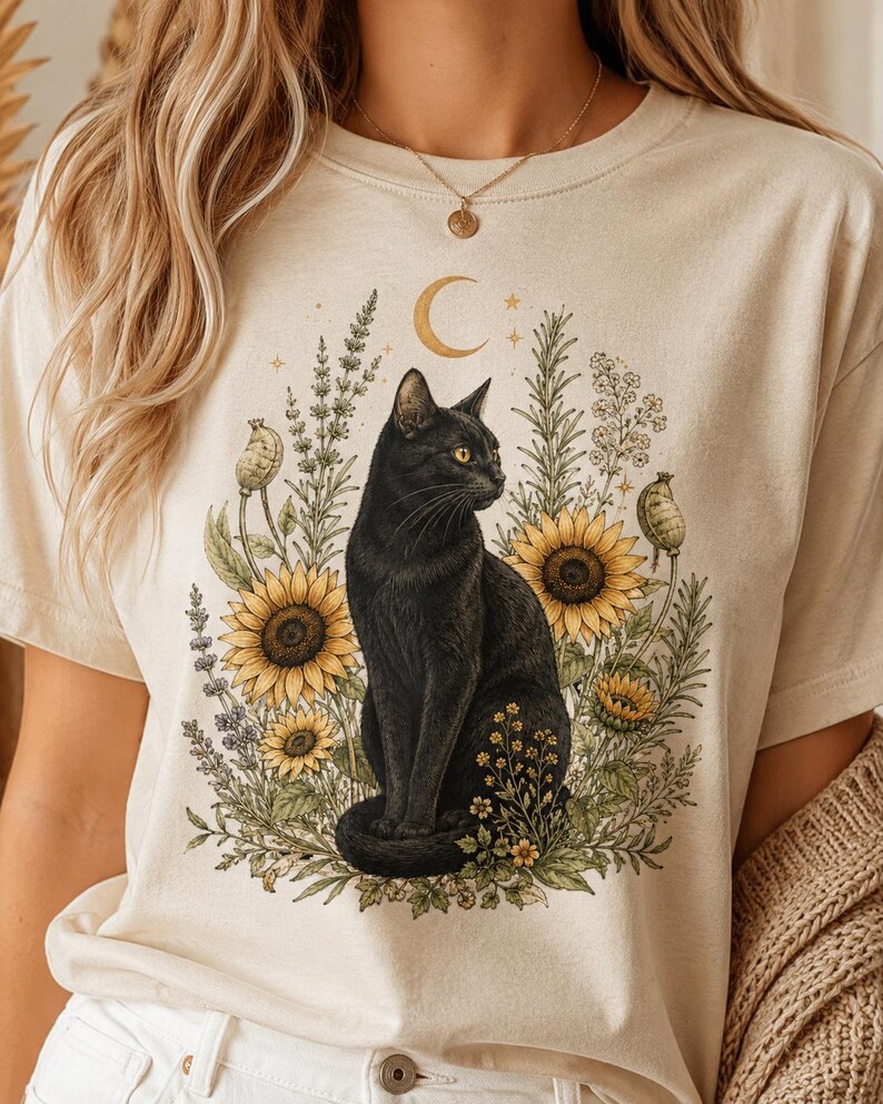 May include: A cream-colored t-shirt featuring a graphic print of a black cat sitting amidst yellow sunflowers, green foliage, and lavender sprigs, topped with a golden crescent moon and small stars, rendered in a detailed, illustrative style.