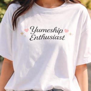 May include: White t-shirt with the text "Yumeship Enthusiast" in a script font, accented with pink hearts and gold stars. The shirt is made of a soft material and has short sleeves.