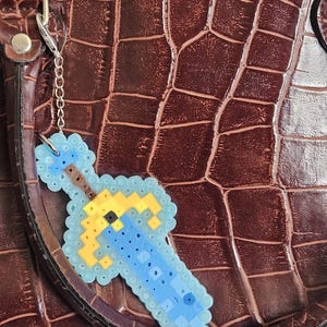 May include: A pixel art sword keychain in shades of blue, yellow, and brown, attached to a silver chain. The keychain is hanging on a brown, textured leather bag. The sword design is made of small, colorful beads.