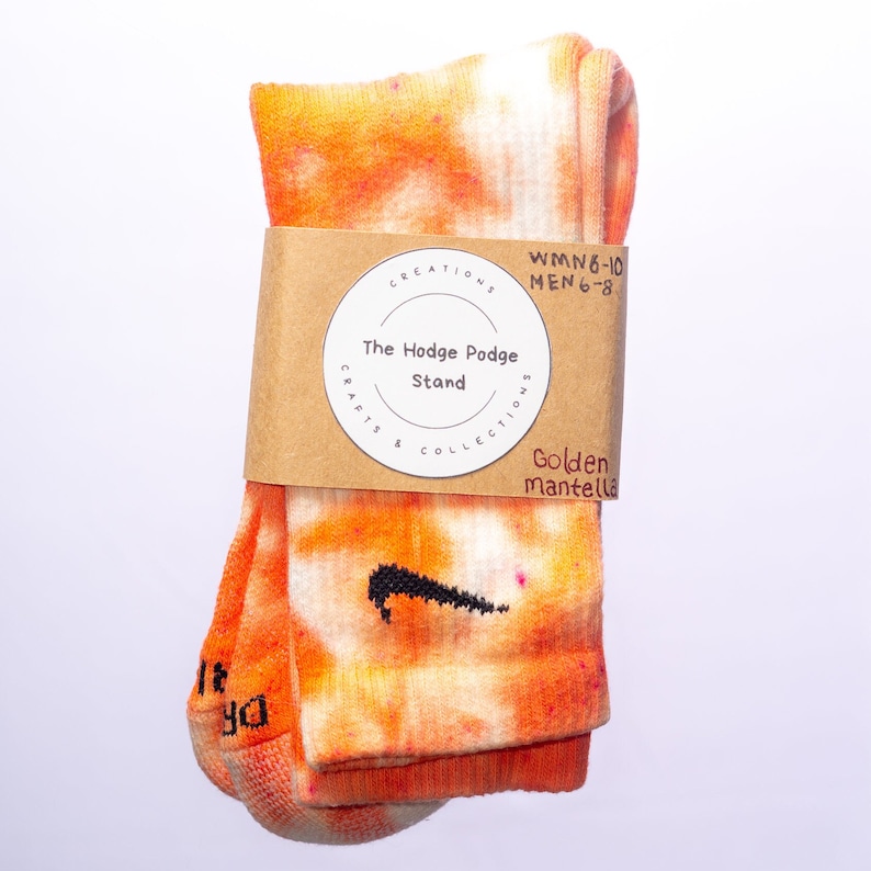 K&ouml;nnte beinhalten: Orangefarbene und wei&szlig;e Tie-Dye-Socken mit einem schwarzen Nike-Swoosh-Logo. Die Socken sind mit einem braunen Papieranh&auml;nger verpackt, auf dem "The Hodge Podge Stand" und "Golden Mantella Creations & Collections" steht.