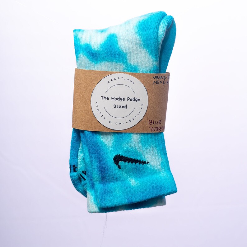 K&ouml;nnte beinhalten: Ein Paar blaue und wei&szlig;e Batik-Socken mit einem schwarzen Nike-Swoosh-Logo. Die Socken sind mit einem braunen Papier-Etikett verpackt, auf dem "The Hodge Podge Stand" und "Blue Dragon" steht.