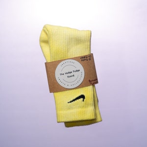 K&ouml;nnte beinhalten: Gelbe Socken mit einem schwarzen Nike-Swoosh-Logo. Die Socken sind gefaltet und haben ein braunes Etikett mit dem Text "The Hodge Podge Stand, Crafts & Collections, Burmese Python, WMN 6-10 MEN 6-8" befestigt.