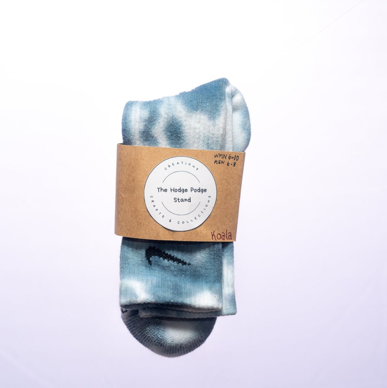 K&ouml;nnte beinhalten: Ein Paar blaue Batik-Socken mit einem schwarzen Nike-Swoosh-Logo. Die Socken sind gefaltet und mit einem braunen Etikett verpackt, auf dem "The Hodge Podge Stand, Crafts & Collections, Koala, WMN 6-10, MEN 6-8" steht.
