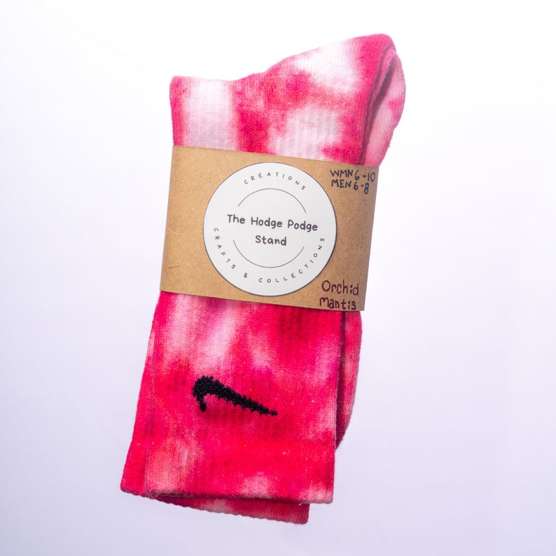 K&ouml;nnte beinhalten: Rosa-wei&szlig;e Batik-Socken mit einem schwarzen Nike-Swoosh-Logo. Die Socken sind gefaltet und haben ein braunes Etikett mit dem Text "The Hodge Podge Stand" und "Orchid Mantis".