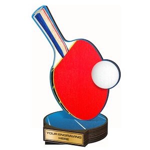 May include: A wooden table tennis trophy with a red paddle, a white ball, and a blue and white base. The base has a gold plaque with the text "YOUR ENGRAVING HERE".