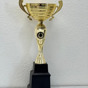 May include: A gold trophy with a black base. The trophy has a cup with two handles and a laurel wreath design on the base.
