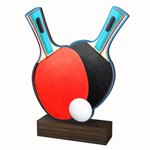 May include: A wooden table tennis trophy with two red and black paddles and a white ball on top. The base is made of dark wood.