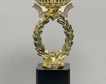 May include: A gold trophy with a laurel wreath design and a black base. The trophy has a cup-shaped top with a decorative rim.