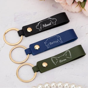 May include: Three personalized leather keychains with gold key rings. The keychains are black, blue, and green. Each keychain has a white outline of a dog breed and a name: Maud, Betsy, and Karen.