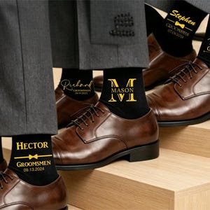 May include: Black socks with gold text and graphics, including names like Stephen, Adam, Richard, Mason, and Hector, along with the title Groomsman or Groomsmen, dates, and bow tie icons. The socks are worn with grey trousers and brown leather dress shoes.