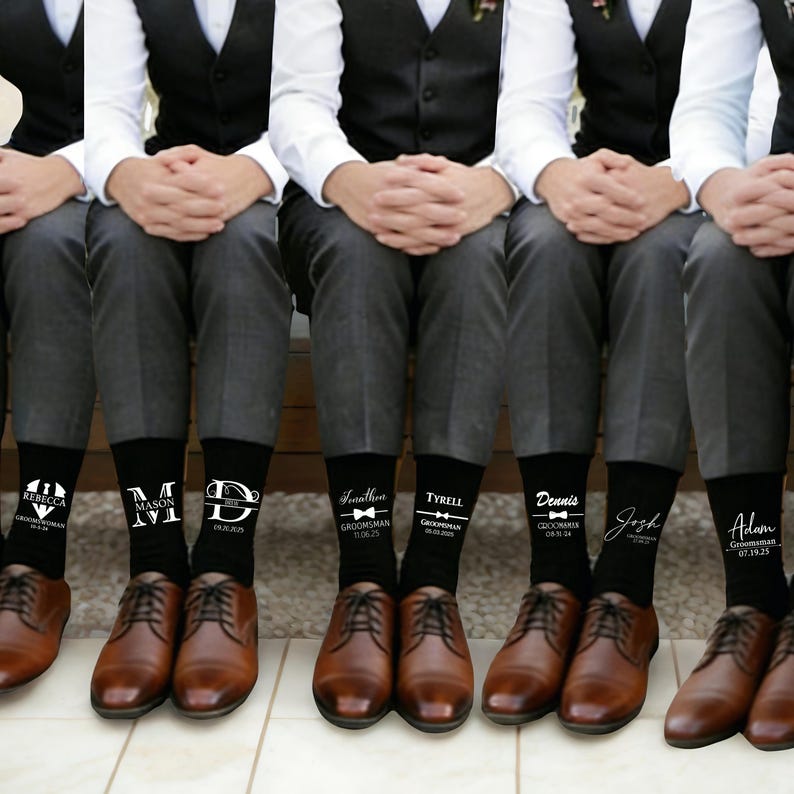 May include: A row of black crew socks worn with grey trousers and brown leather dress shoes. Each sock features white custom text and graphics, including names like Rebecca, Mason, Jonathan, Tyrell, Dennis, Josh, and Adam, along with dates.