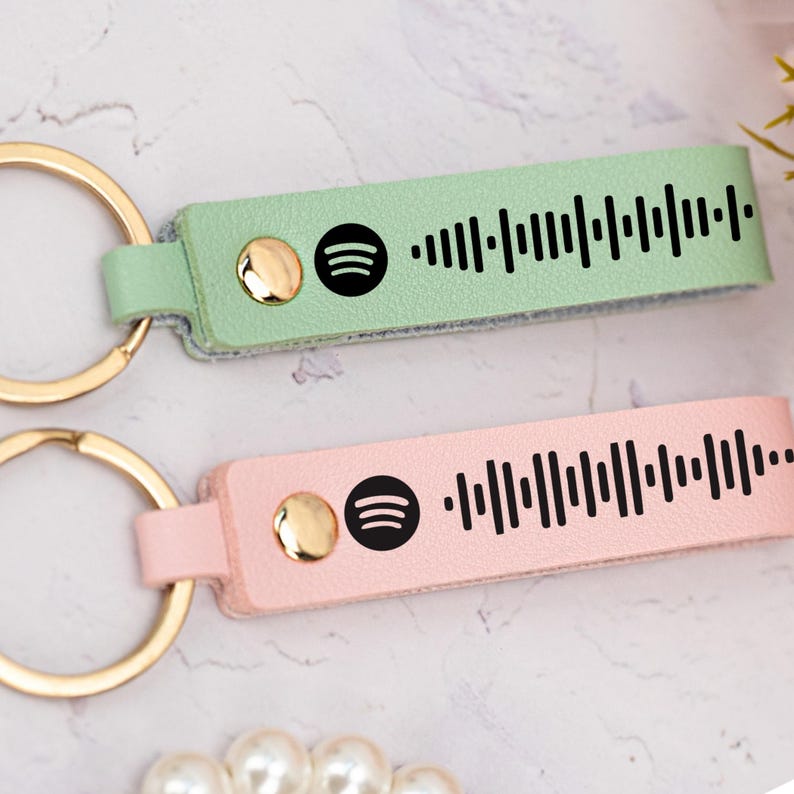 May include: Two leather keychains with gold key rings. One keychain is light green with a black Spotify logo and a black barcode. The other keychain is pink with a black Spotify logo and a black barcode.