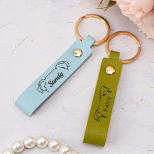 May include: Two personalized leather keychains with gold key rings. The blue keychain has a dog silhouette and the name "Sandy" engraved on it. The green keychain has a mountain silhouette and the name "Fairy" engraved on it.