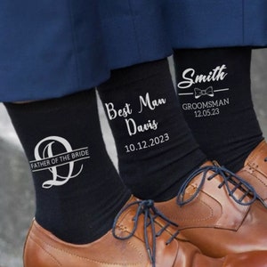 May include: Three pairs of black socks with white text, worn with brown leather dress shoes. One sock reads "Father of the Bride", another "Best Man Davis 10.12.2023", and the third "Smith GROOMSMAN 12.05.23". The socks are visible from the ankles down.