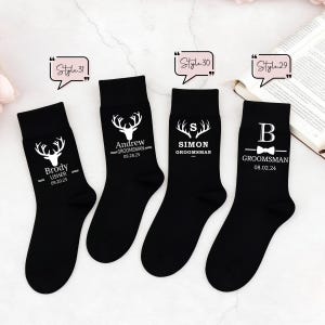 May include: Four black crew socks featuring white printed designs. Each sock displays a unique graphic, including deer antlers, names like Brody, Andrew, and Simon, titles such as Groomsman or Usher, dates, and the text Style 29, 30, and 31 in speech bubbles.