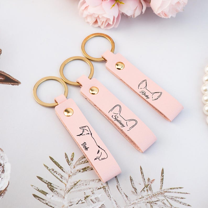May include: Three pink leather keychains with gold key rings. Each keychain has a personalized name and a line drawing of a dog breed. The names are "Bob", "Sydney", and "Ricky".