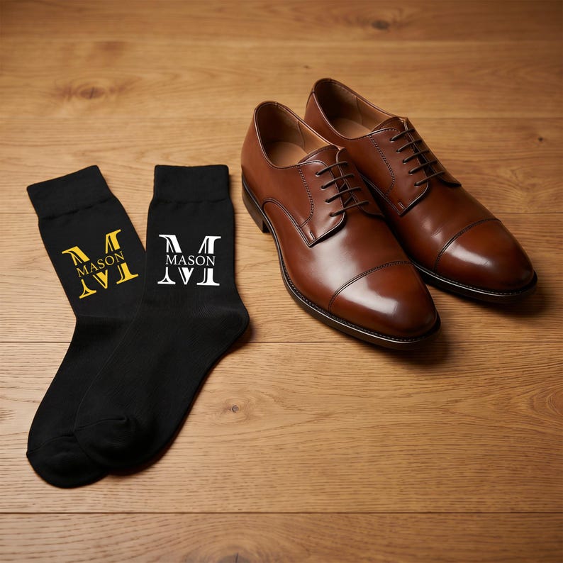 May include: A pair of black socks and a pair of brown leather dress shoes. One sock features a yellow letter M with the text MASON, while the other sock features a white letter M with the text MASON. The shoes have laces and a cap toe design.