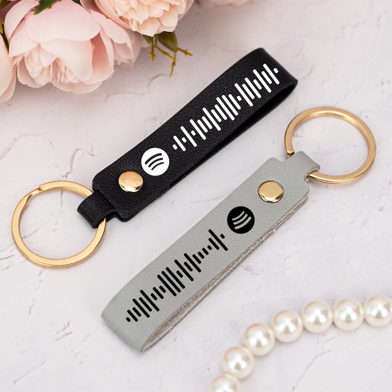 May include: Two leather keychains with gold key rings. One keychain is black with a white Spotify logo and barcode. The other keychain is gray with a black Spotify logo and barcode.
