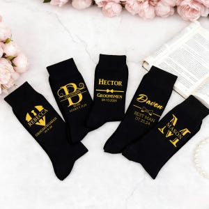 May include: Five black socks featuring gold printed text and graphics. The socks display names including Rebecca, Bryan, Hector, Davon, and Mason, along with titles like Groomswoman, Groomsman, and Best Man, plus specific dates and bow tie icons.