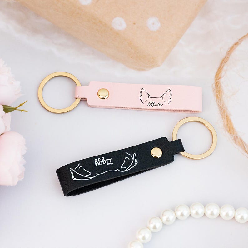 May include: Two personalized leather keychains with gold rings. The pink keychain has a dog silhouette and the text "Rocky". The black keychain has a dog paw print and the text "Hobbrz".