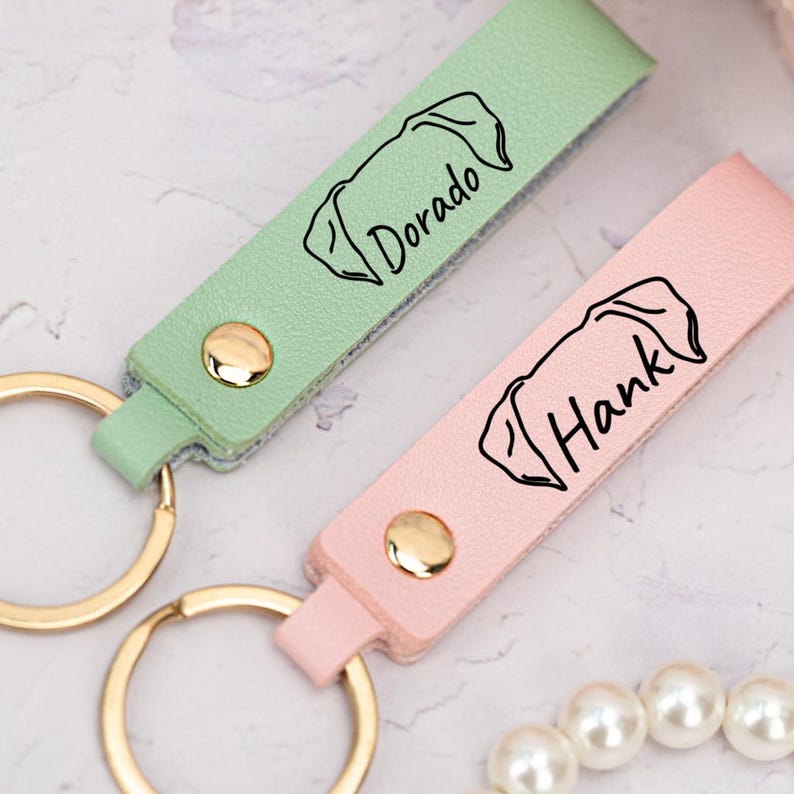 May include: Two leather keychains with gold rings. One is light green with the name "Dorado" and the other is pink with the name "Hank". Both have a dog silhouette engraved on them.