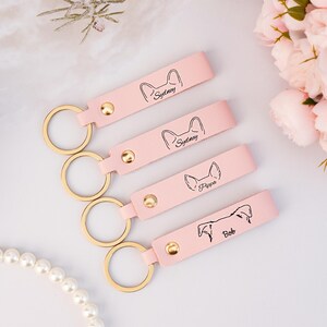 May include: Four pink leather keychains with gold key rings. Each keychain has a different dog breed silhouette and the name of the dog: Sydney, Sydney, Pippa, and Bob.