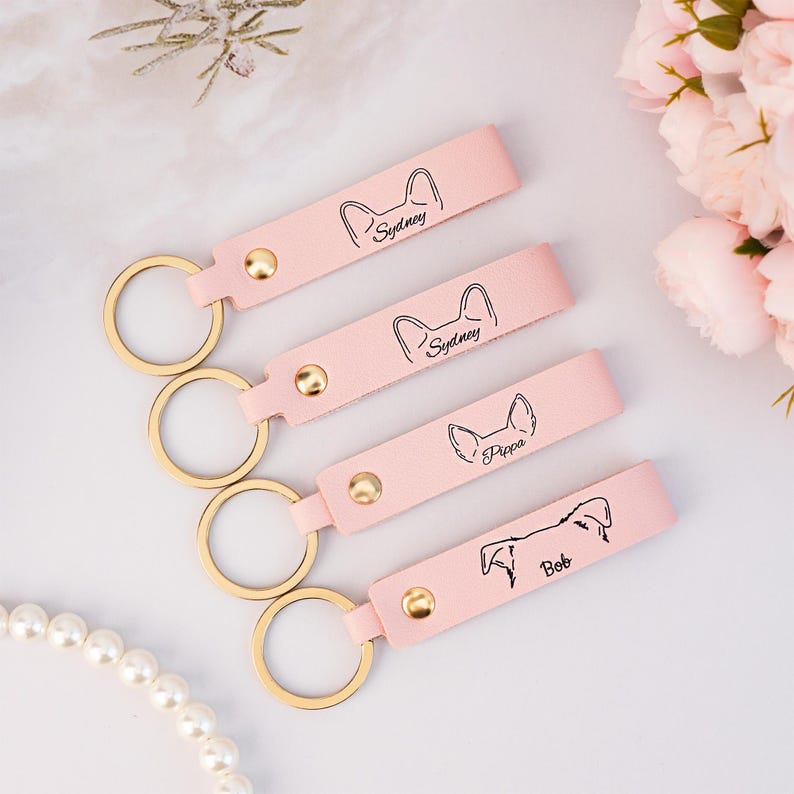 May include: Four pink leather keychains with gold key rings. Each keychain has a different dog breed silhouette and the name of the dog: Sydney, Sydney, Pippa, and Bob.
