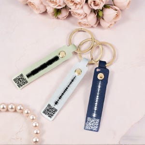 May include: Three leather keyrings in pastel green, light blue, and navy blue, each with a gold-coloured key ring. The keyrings feature a sound wave design and a QR code. A gold snap secures the leather strap to the key ring.