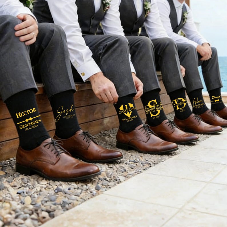 May include: A row of black dress socks worn with brown leather shoes. Each sock features custom gold text and graphics, including names like Hector, Josh, Cameron, Shohilbhai, Adam, and David, along with the title Groomsman and wedding dates.