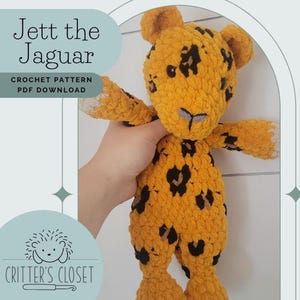 Pode incluir: Um brinquedo de pelúcia de crochê em forma de onça-pintada chamado Jett. O brinquedo é amarelo com manchas pretas e tem um nariz roxo claro. O brinquedo está sendo segurado por uma mão. O texto "Jett the Jaguar" e "Crochet Pattern PDF Download" é visível na imagem. A imagem também inclui um logotipo para "Critter's Closet".