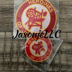 May include: Two embroidered patches in clear plastic packaging. The larger patch is circular with a red border and the words "HEROINES OF" in white. The center has a red illustration. The smaller patch has the words "HEROINES OF JERICHO".