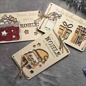 K&ouml;nnte beinhalten: Geschenkkartenhalter aus Holz mit ausgeschnittenen Designs und deutschem Text. Einer zeigt ein Santa-Design, ein anderer eine Geschenkt&uuml;te und der dritte Geschenkboxen. Jedes ist mit Bindfaden gebunden. Der Text enth&auml;lt "Frohe Weihnachten" und "Dieses Jahr gibt es KOHLE".