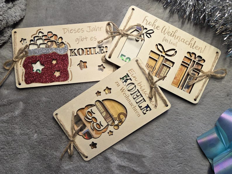 K&ouml;nnte beinhalten: Geschenkkartenhalter aus Holz mit ausgeschnittenen Designs und deutschem Text. Einer zeigt ein Santa-Design, ein anderer eine Geschenkt&uuml;te und der dritte Geschenkboxen. Jedes ist mit Bindfaden gebunden. Der Text enth&auml;lt "Frohe Weihnachten" und "Dieses Jahr gibt es KOHLE".