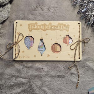 K&ouml;nnte beinhalten: Weihnachtskarte aus Holz mit ausgeschnittenen Ornamentformen, die Geldscheine zeigen. Aufschrift "Frohe Weihnachten" und "f&uuml;r Katrin". Goldene Schnur und Schleifen zur Dekoration. Die Karte ist ca. 15 cm lang.